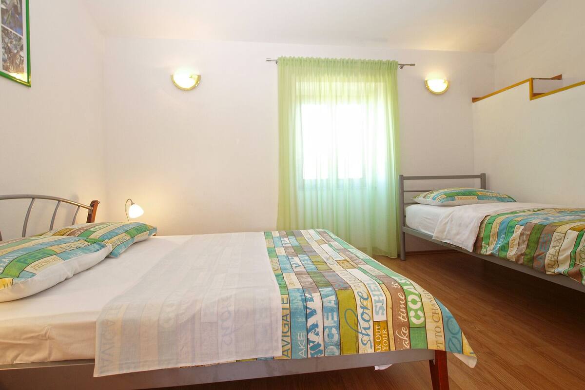 A room with two beds, a window with green curtains, and wall-mounted lights.