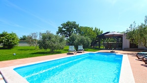 A rectangular swimming pool with a tiled edge, surrounded by a paved area. There are two lounge chairs with floral upholstery and a covered seating area in the background. - House 1319 (Liznjan)
