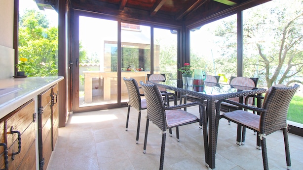 A covered patio with a glass table and wicker chairs, overlooking a garden and a building. - House 1319 (Liznjan)