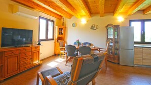 A cozy cabin interior with wooden beams, a dining area with chairs, a kitchen with a refrigerator, and a television on a wooden cabinet. - House 1319 (Liznjan)