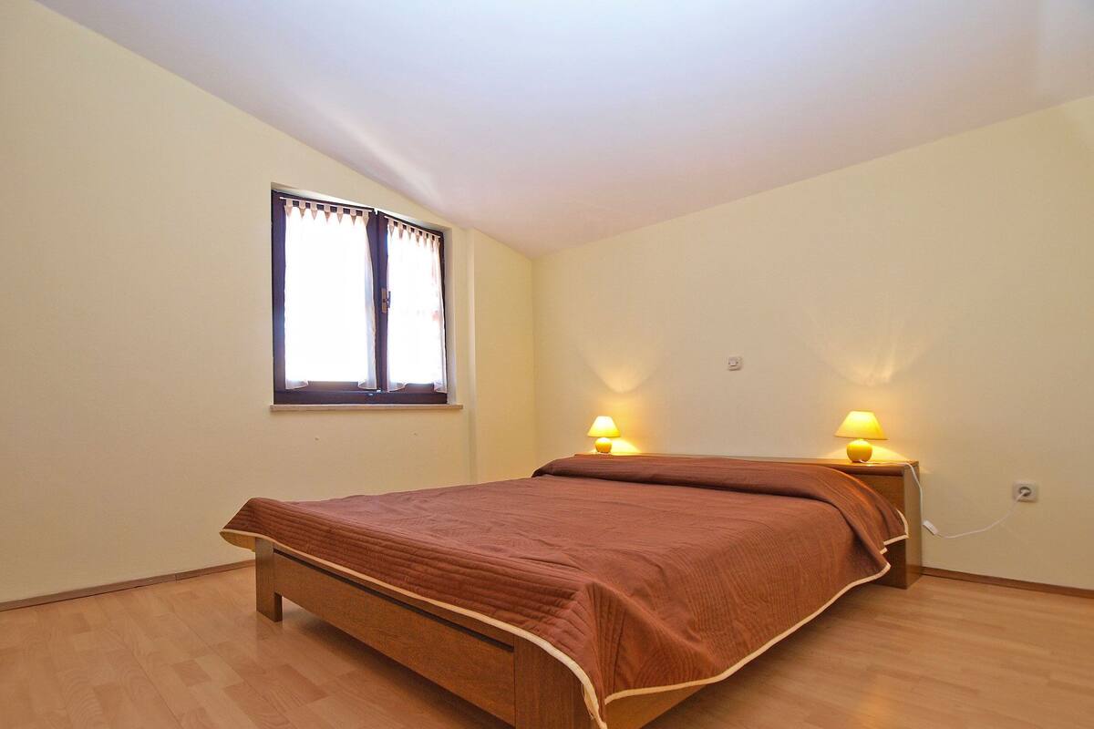 Un dormitorio con una cama de madera, dos lámparas de noche, un ventanal con cortinas y un interruptor de luz en la pared.