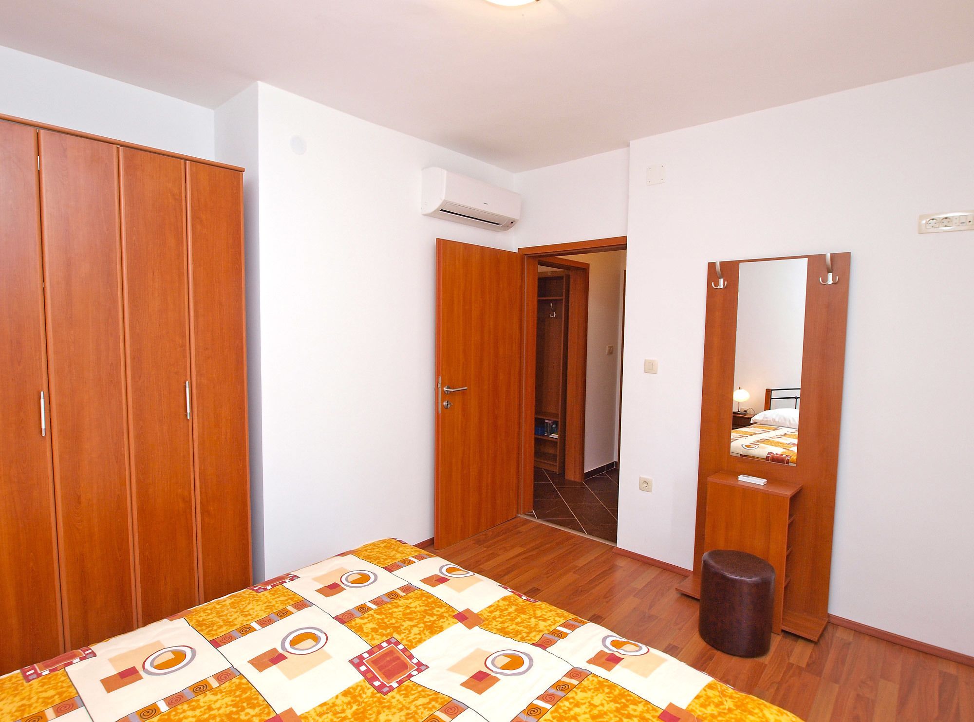 Ein Schlafzimmer mit Holzschrank, Bett mit gemustertem Überwurf, Holzschemel und Spiegel an der Wand.