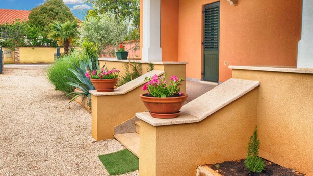 Ein Haus mit einer beige farbenen Fassade, einem roten Ziegeldach und einem kleinen Garten mit Topfpflanzen und einem grünen Rasen.