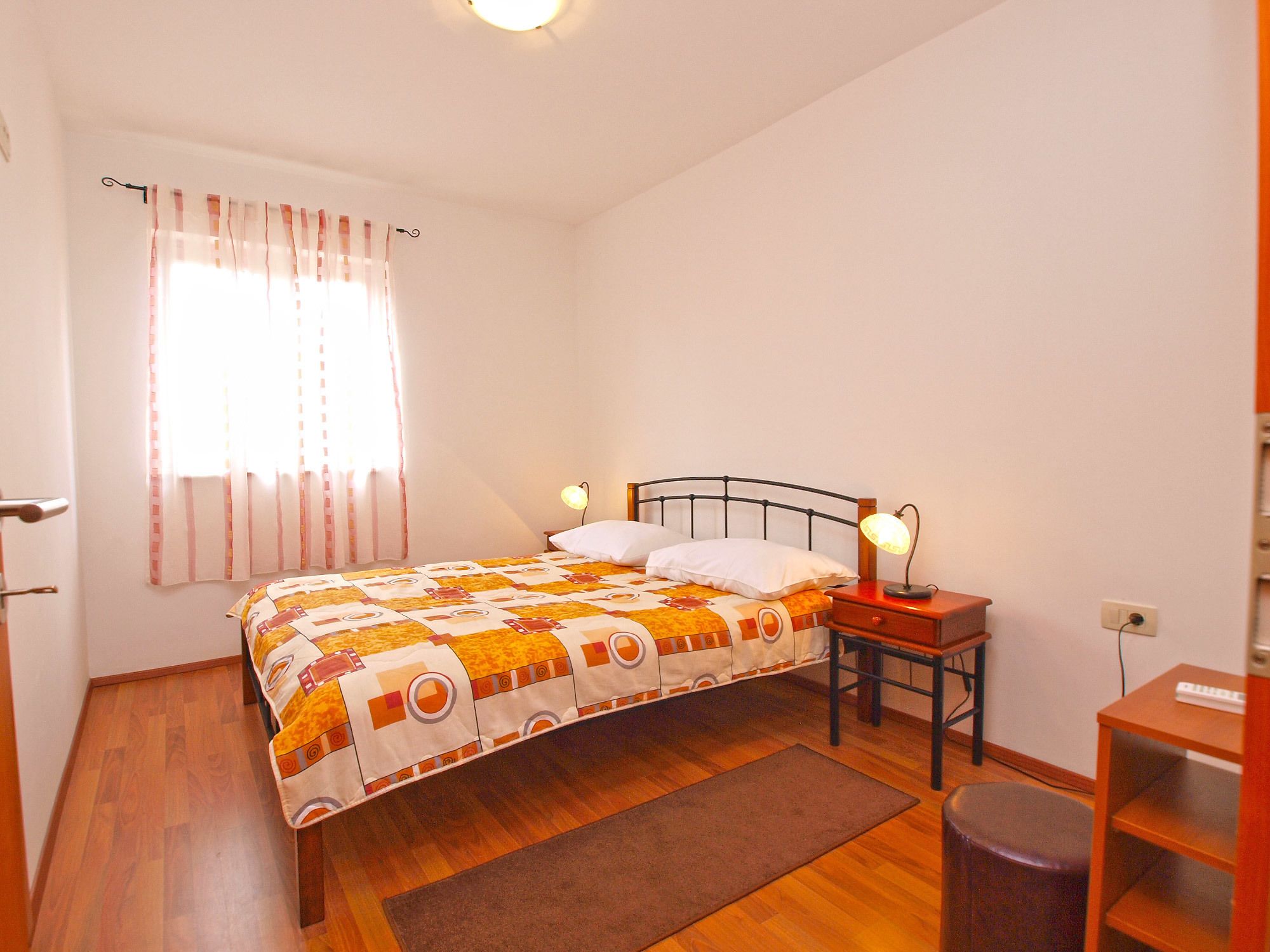 A bedroom with a bed, bedside lamps, a nightstand, a wooden floor, a window with curtains, and a wall socket.