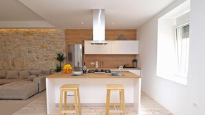 A modern kitchen with a central island, two wooden stools, a stone wall, a wooden ceiling, and a large window. - House 1338 (Pula)