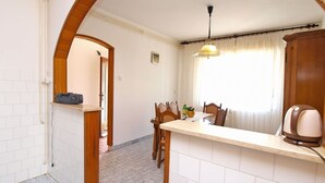 A compact kitchen with a dining area, featuring a tiled backsplash, wooden chairs, a small table, and a window with curtains. - Apartment 1341 (Pula)