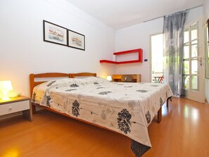 A bedroom with a wooden bed, a bedside table with a lamp, a red shelf, and two framed pictures on the wall. - Apartment 1346 (Pula)