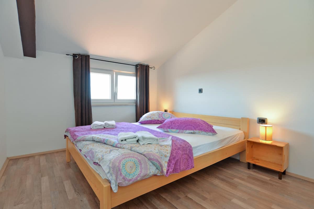A bedroom with a wooden bed, two bedside tables with lamps, a window with curtains, and a wooden floor.