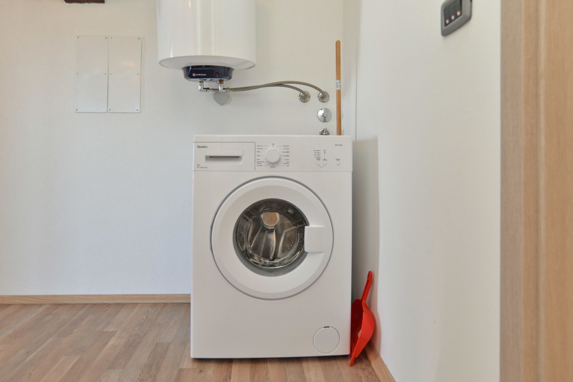 Een compacte, witte wasmachine in een kleine wasruimte met een houten vloer.