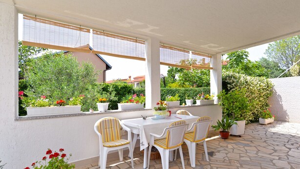 A covered patio with a table and chairs, surrounded by potted plants and a view of greenery. - Apartment 1375 (Fazana)