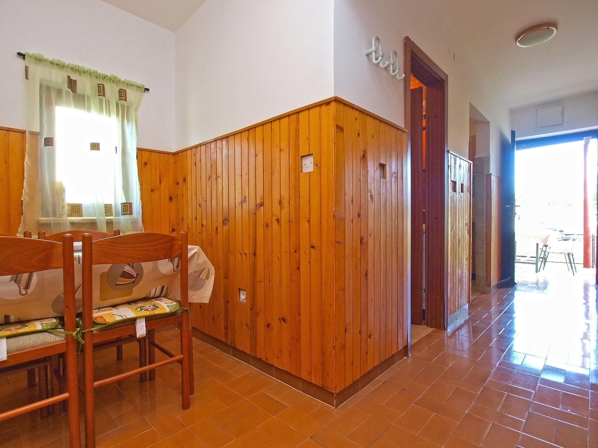A room with wooden paneling, a wooden chair, a window with curtains, and a door leading to another room.