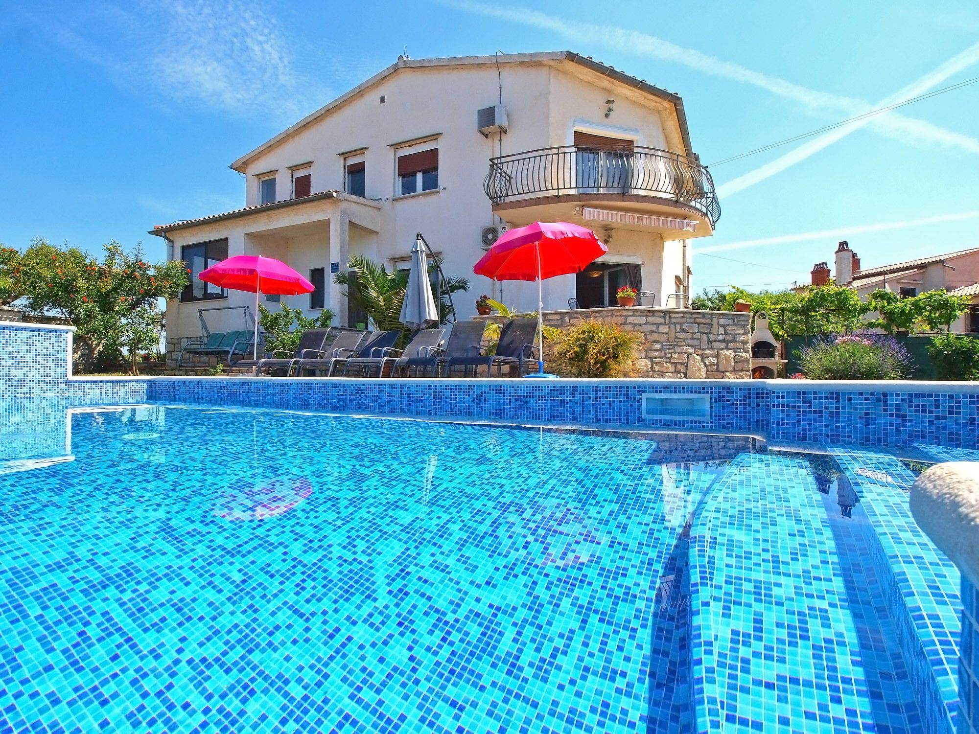 A swimming pool with blue mosaic tiles, surrounded by lounge chairs and red umbrellas.