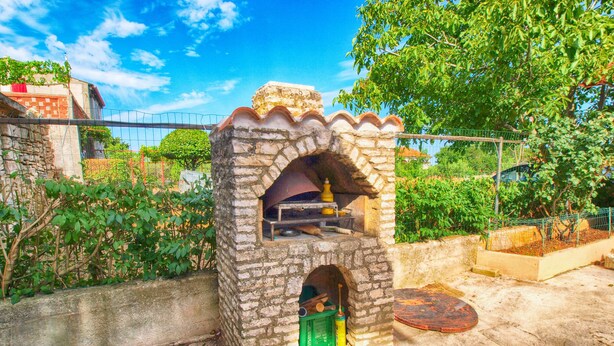 A brick-built outdoor oven with a chimney, surrounded by greenery and a fence. - Apartment 1388 (Vodnjan)