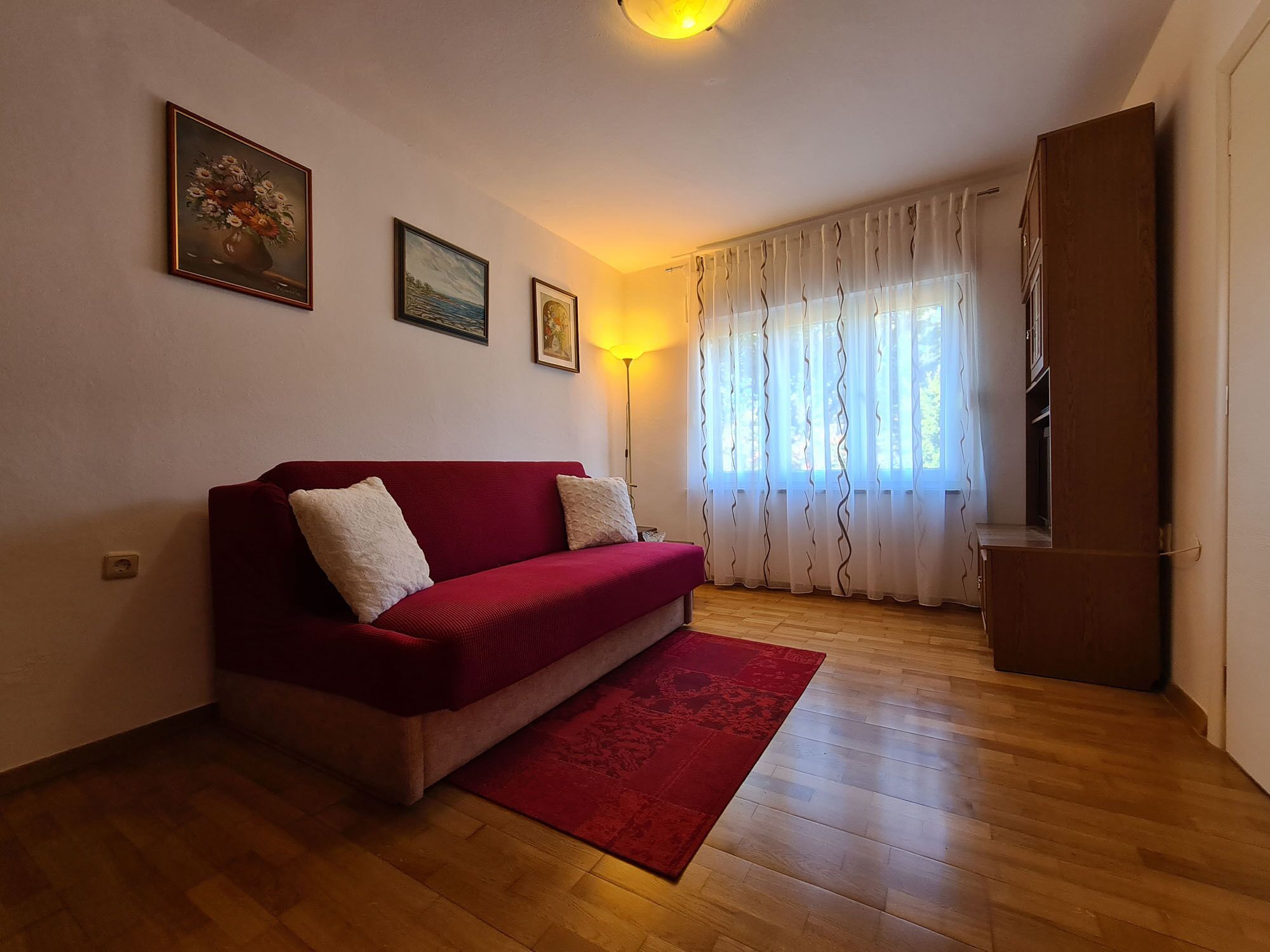 Un salon avec un canapé rouge, un tapis rouge, un plancher en bois et une fenêtre avec des rideaux.