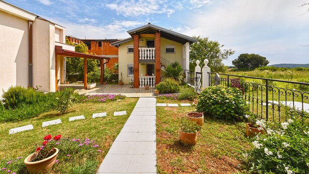 Une maison à deux étages dotée d’un patio couvert, d’un jardin agrémenté de plantes en pot et d’un chemin menant à l’entrée.