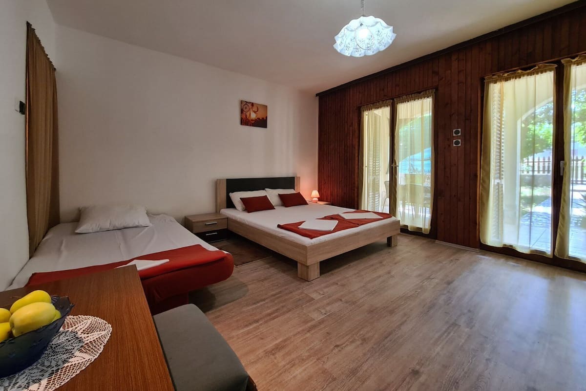 A bedroom with two beds, a wooden floor, a desk with a bowl of fruit, and a window with curtains.