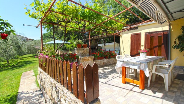 Un área de estar al aire libre con techo, con una mesa y sillas, rodeada por un muro de piedra y una valla de madera.