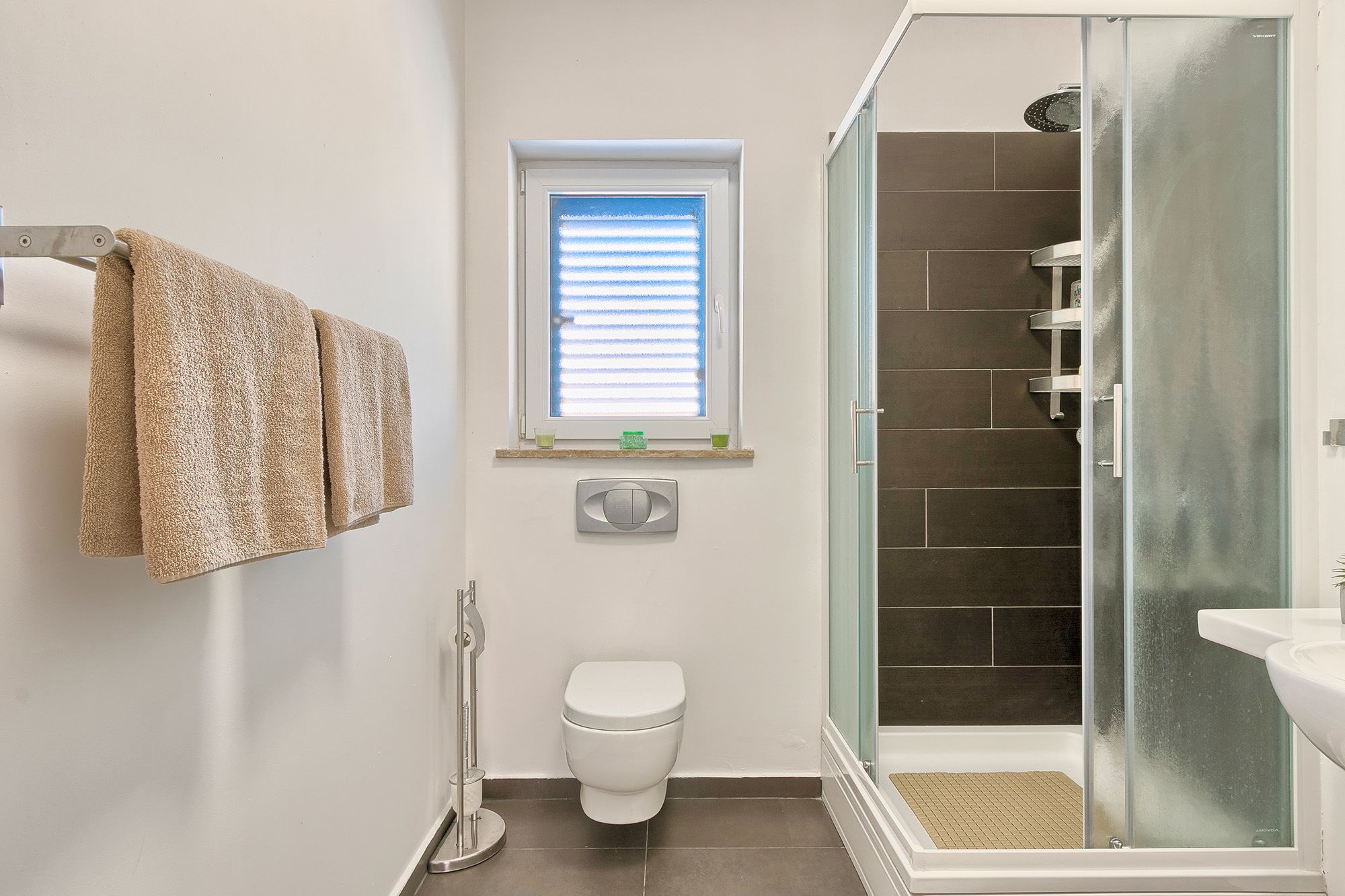 A bathroom with a toilet, a window with blinds, a shower with a glass door, and a towel rack with towels.