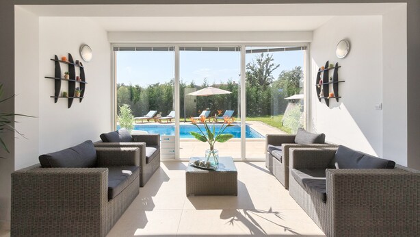 Ein sonnendurchflutetes Zimmer mit Korbmöbeln, einem Glastisch und Blick auf einen Swimmingpool.