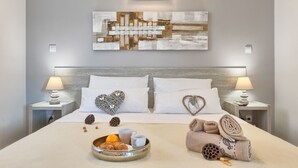 Ein ordentlich bezogenes Bett mit weißen Laken, zwei Nachttischlampen, dekorativen Kissen, einem Wandbild und einem Nachttisch mit einer Obstschale und einer Tasse.