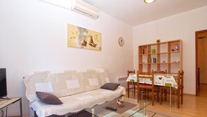 A living room with a sofa, a glass coffee table, a dining table with chairs, a bookshelf, and an air conditioning unit. - Apartment 1407 (Fazana)