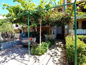 A car parked in a covered parking area with a pergola overhead, a building with a white facade, and a staircase leading to a door. - Apartment 1407 (Fazana)