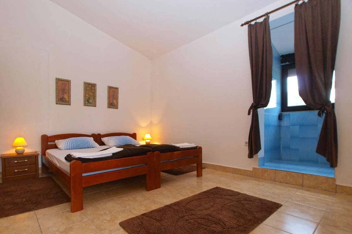 A bedroom with a double bed, a nightstand with a lamp, a brown rug, and a window with curtains.