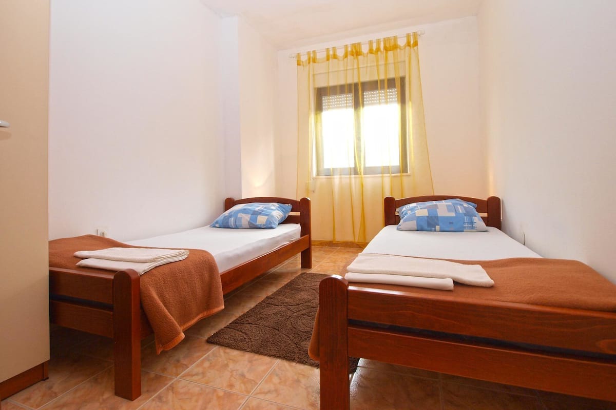 A small, clean room with two single beds, a window with yellow curtains, and a brown rug on the floor.