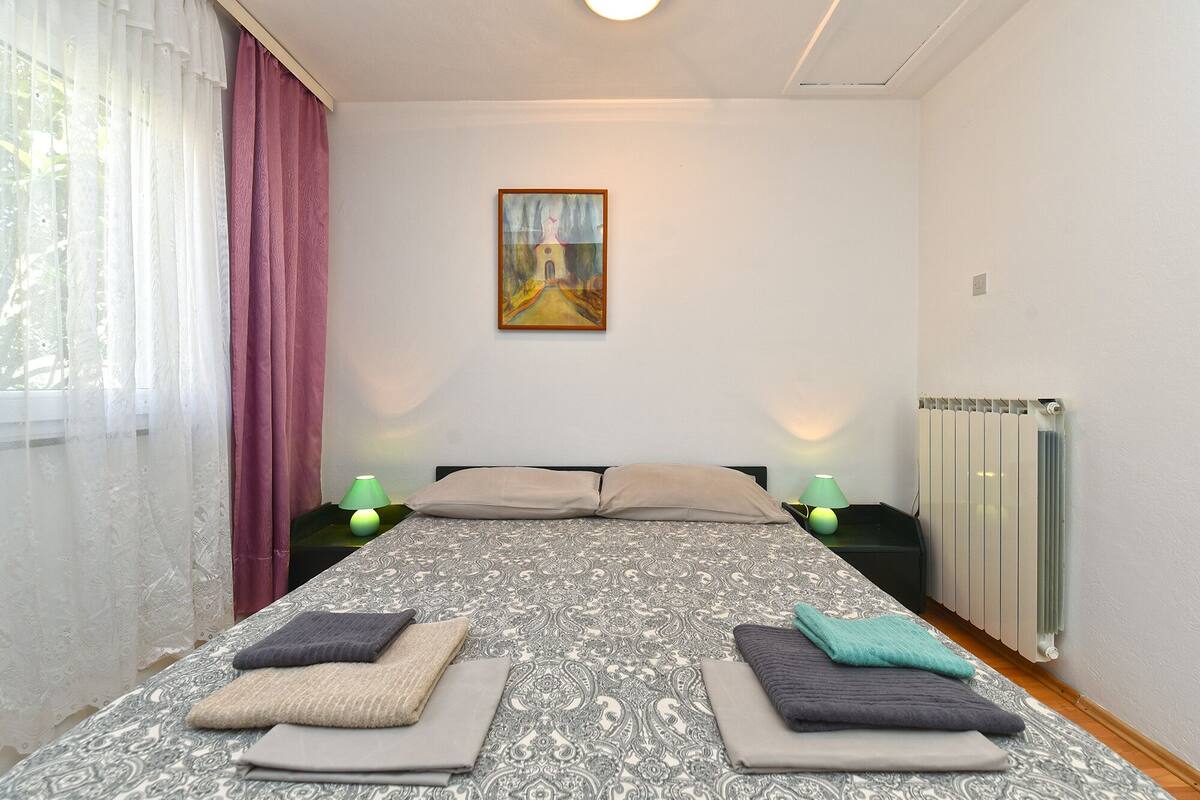 A bedroom with a bed, two bedside tables, a radiator, and a painting on the wall.