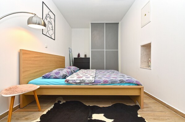 Quarto com cama de madeira, um pequeno banquinho, tapete preto e branco, luminária na parede e um quadro emoldurado.