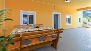 A room with a wooden bench, a table with a checkered tablecloth, and two windows with curtains. - House 1411 (Marcana)