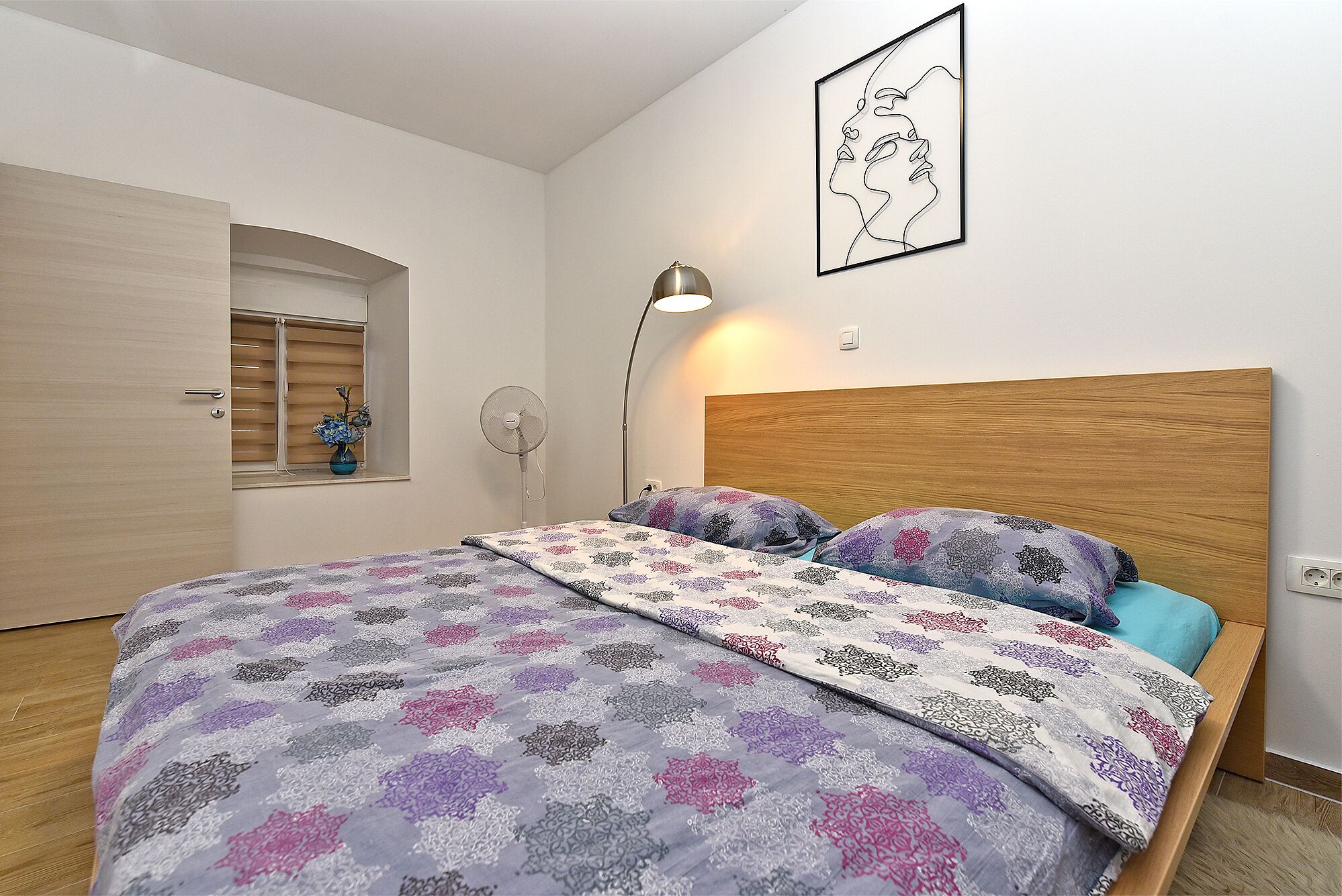 A bedroom with a wooden headboard, a bed with patterned bedding, a bedside lamp, and a wall-mounted artwork.