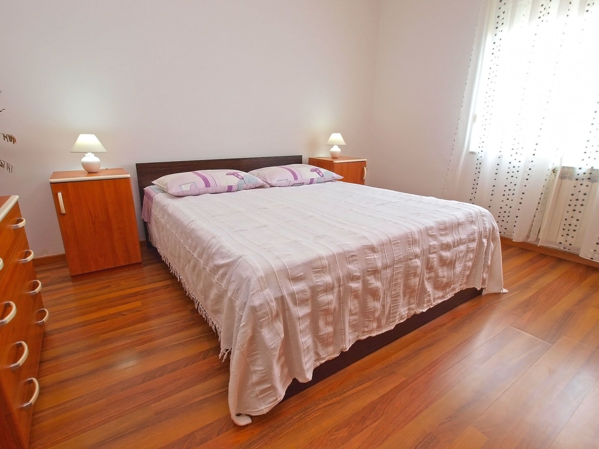 A bedroom with a wooden floor, a bed with a white bedspread, two bedside tables with lamps, and a window with curtains.