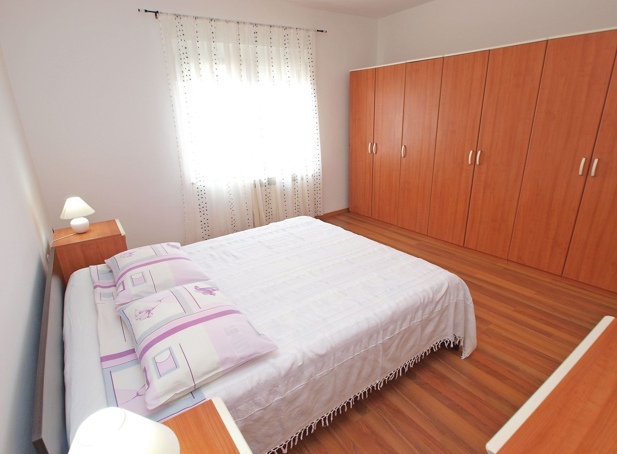 A bedroom with a bed, wooden wardrobe, and a nightstand with a lamp.