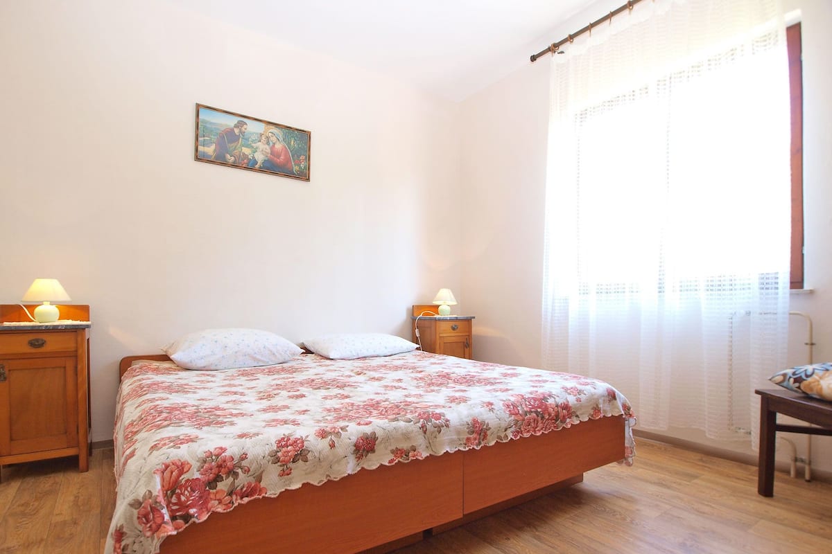 Une chambre à coucher avec un lit en bois, deux tables de chevet avec des lampes, une commode, une fenêtre avec des rideaux et un tableau accroché au mur.