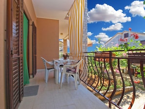 A balcony with a table and chairs, a striped awning, and a view of a garden with flowers and a building in the background.