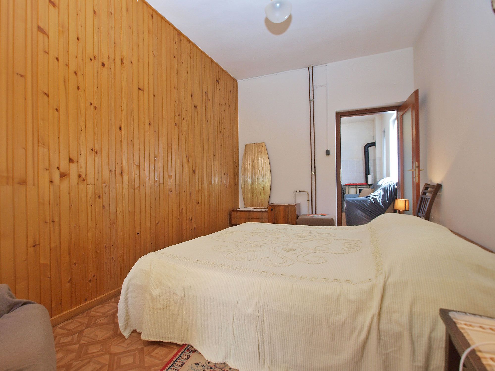 A bedroom with a wooden panel wall, a bed with a white bedspread, a nightstand, a lamp, and a mirror.