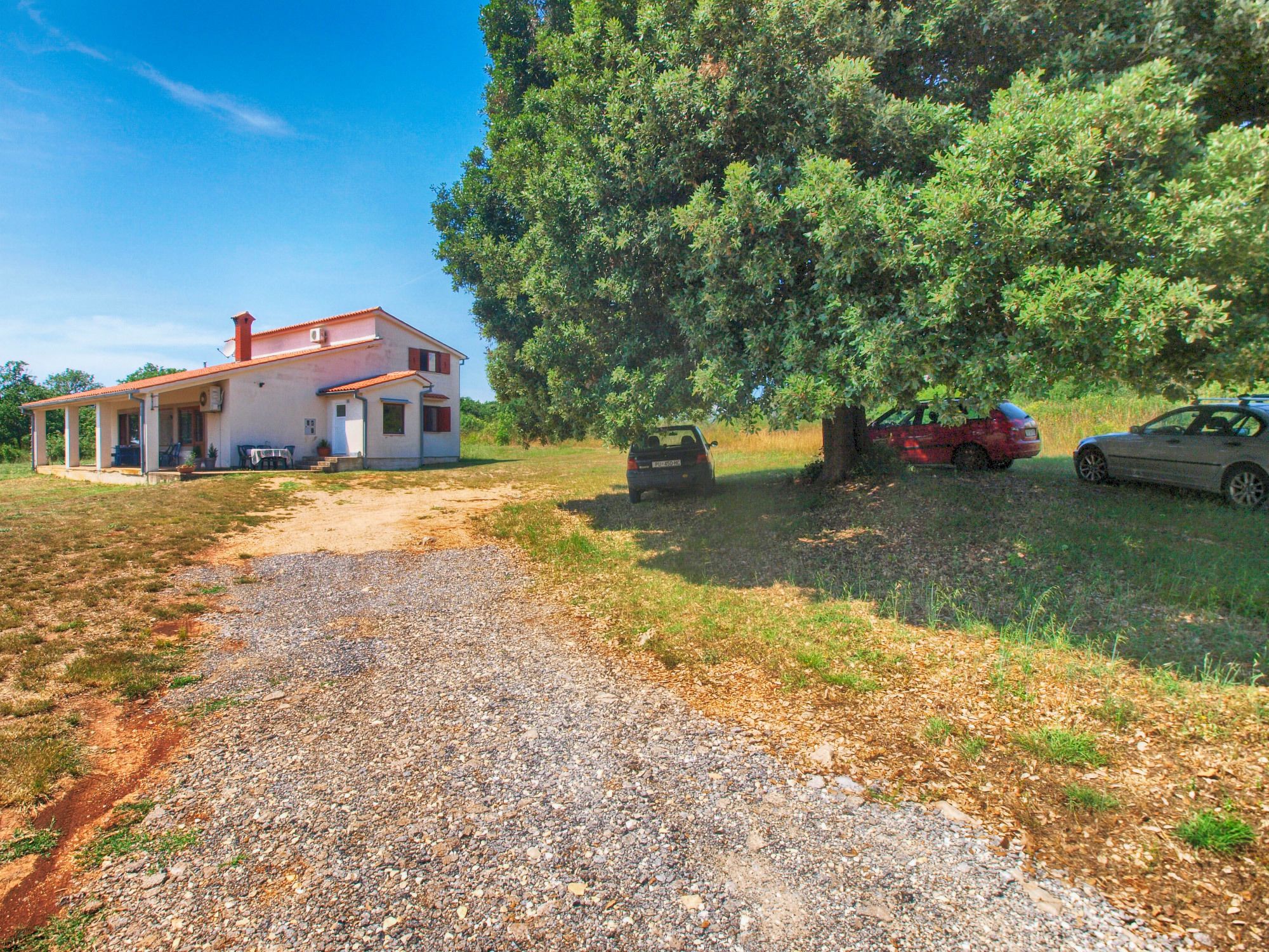 A house with a gravel driveway, a covered porch, and a large tree in the background.
