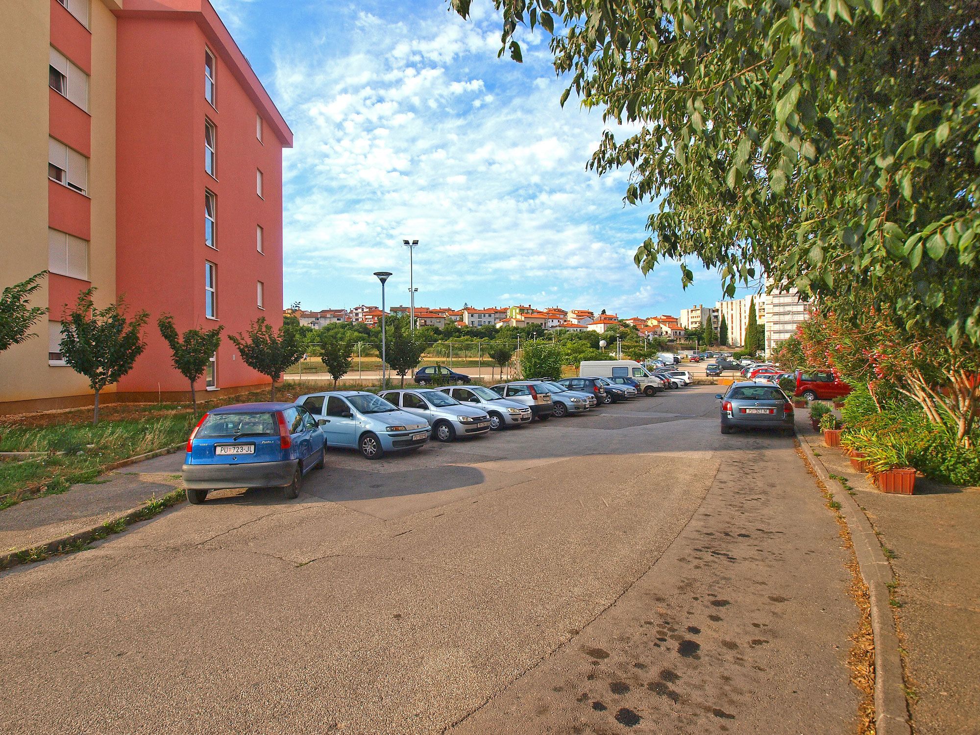 Un estacionamiento con varios coches aparcados en filas, un edificio rojo a la izquierda y una fila de árboles a la derecha.