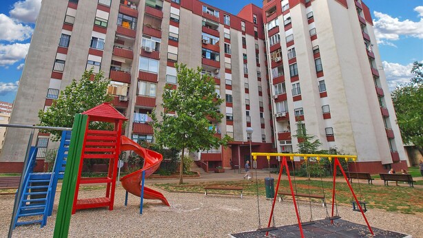 A playground with a red slide and swings, surrounded by a building with multiple balconies and a clear sky. - Apartment 1431 (Pula)