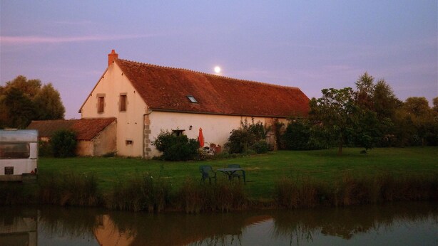 Un bâtiment blanc avec un toit rouge, un étang devant et une lune dans le ciel.