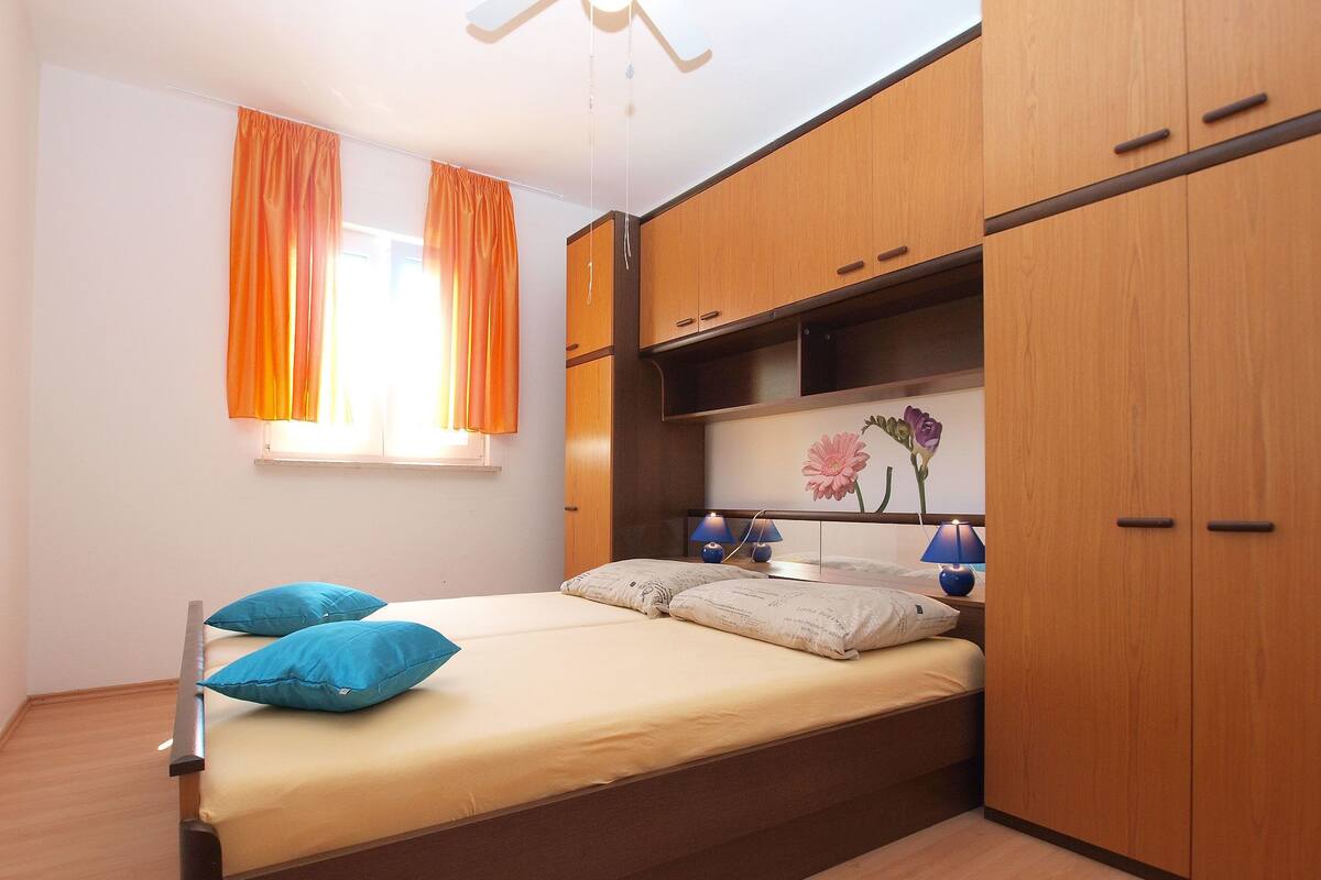 A bedroom with a bed, two pillows, a nightstand with lamps, a wardrobe, and a window with curtains.