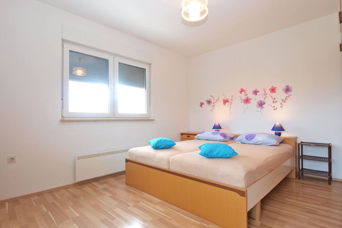 A bedroom with a wooden bed, two bedside tables with lamps, a window with blinds, and a wall with floral decorations.