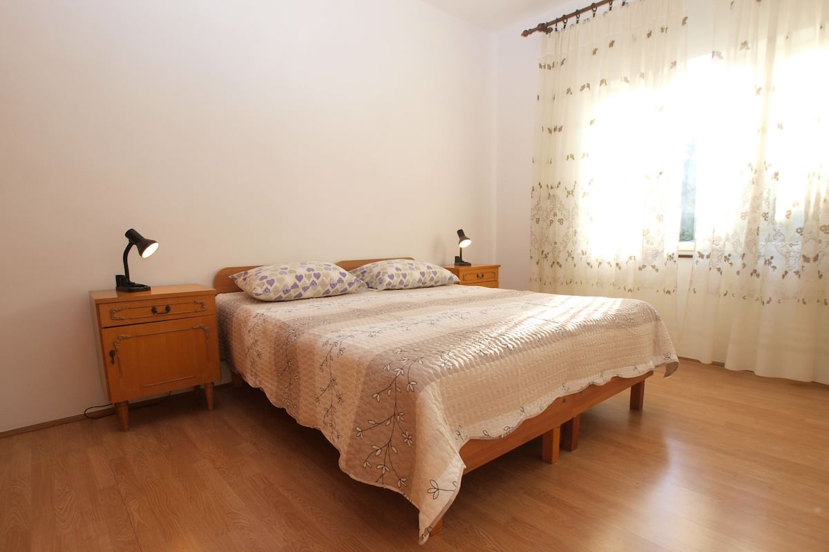Une chambre à coucher avec un lit en bois, deux tables de chevet avec des lampes, une couvre-lit à motifs floraux et des rideaux légers ornés de motifs floraux.