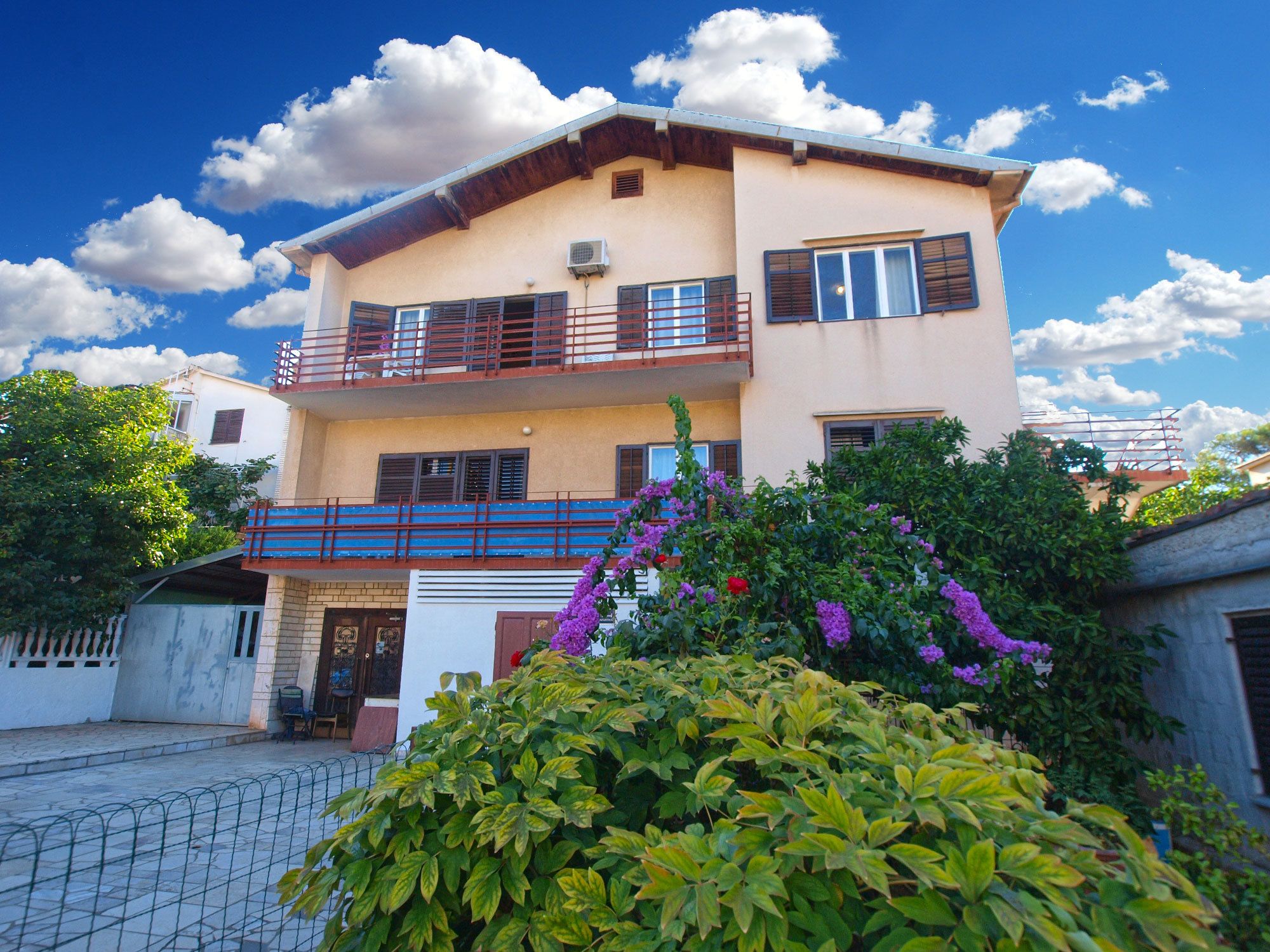 A two-story building with a balcony, a garage, and a garden with purple flowers.