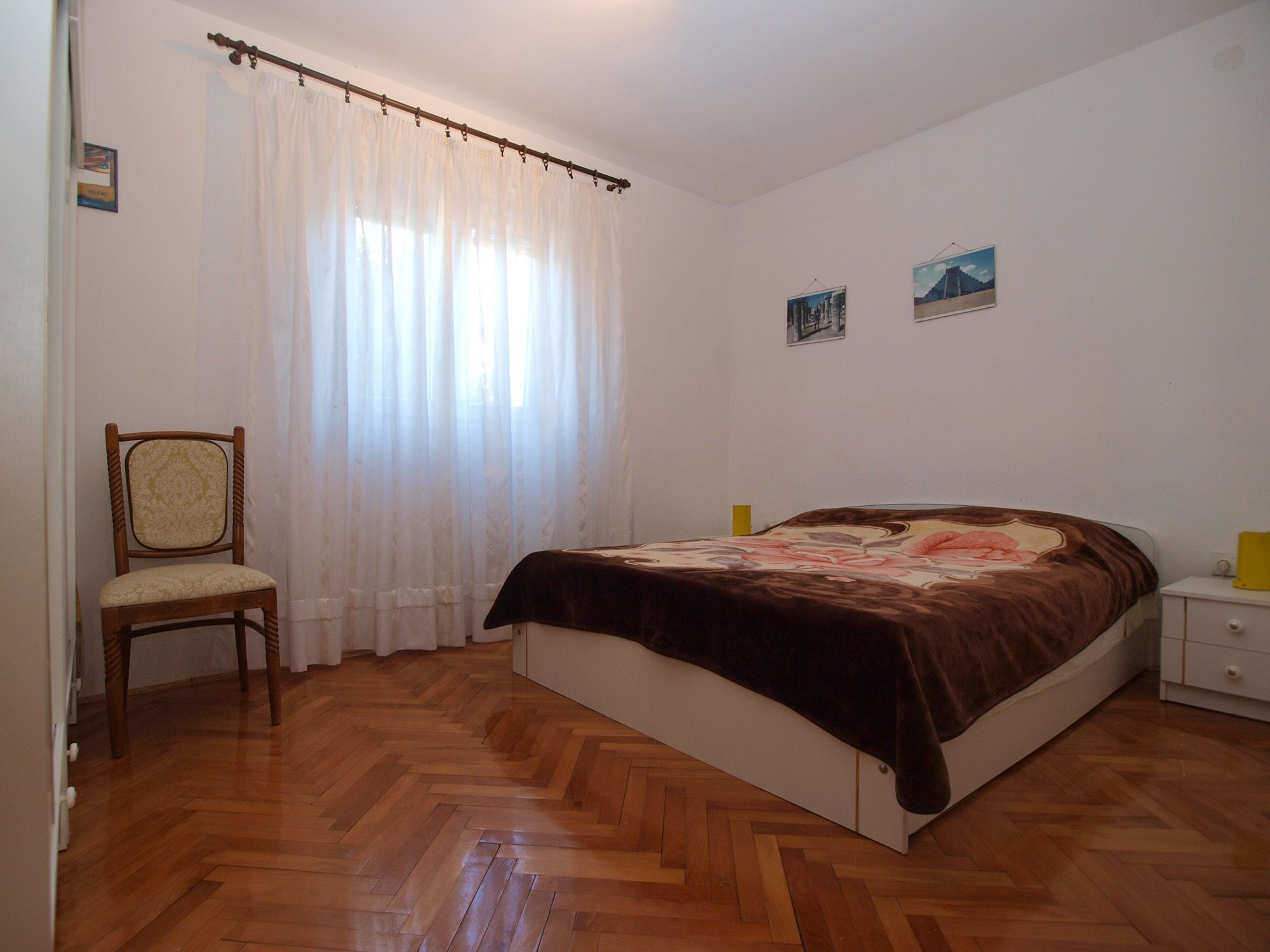 Un dormitorio con piso de madera en patrón espiga, cama con cubrecama floral, una silla, una mesita de noche y una ventana con cortinas de encaje.
