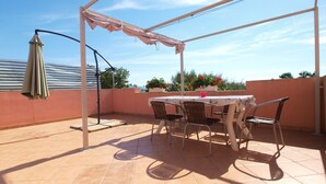Een overdekt terras met een tafel, stoelen, een hangende parasol en potplanten.