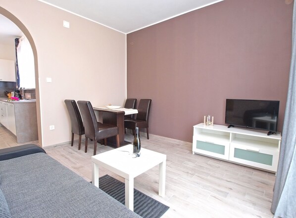 A modern living room with a dining area, a flat-screen TV, and a kitchen visible through an archway.