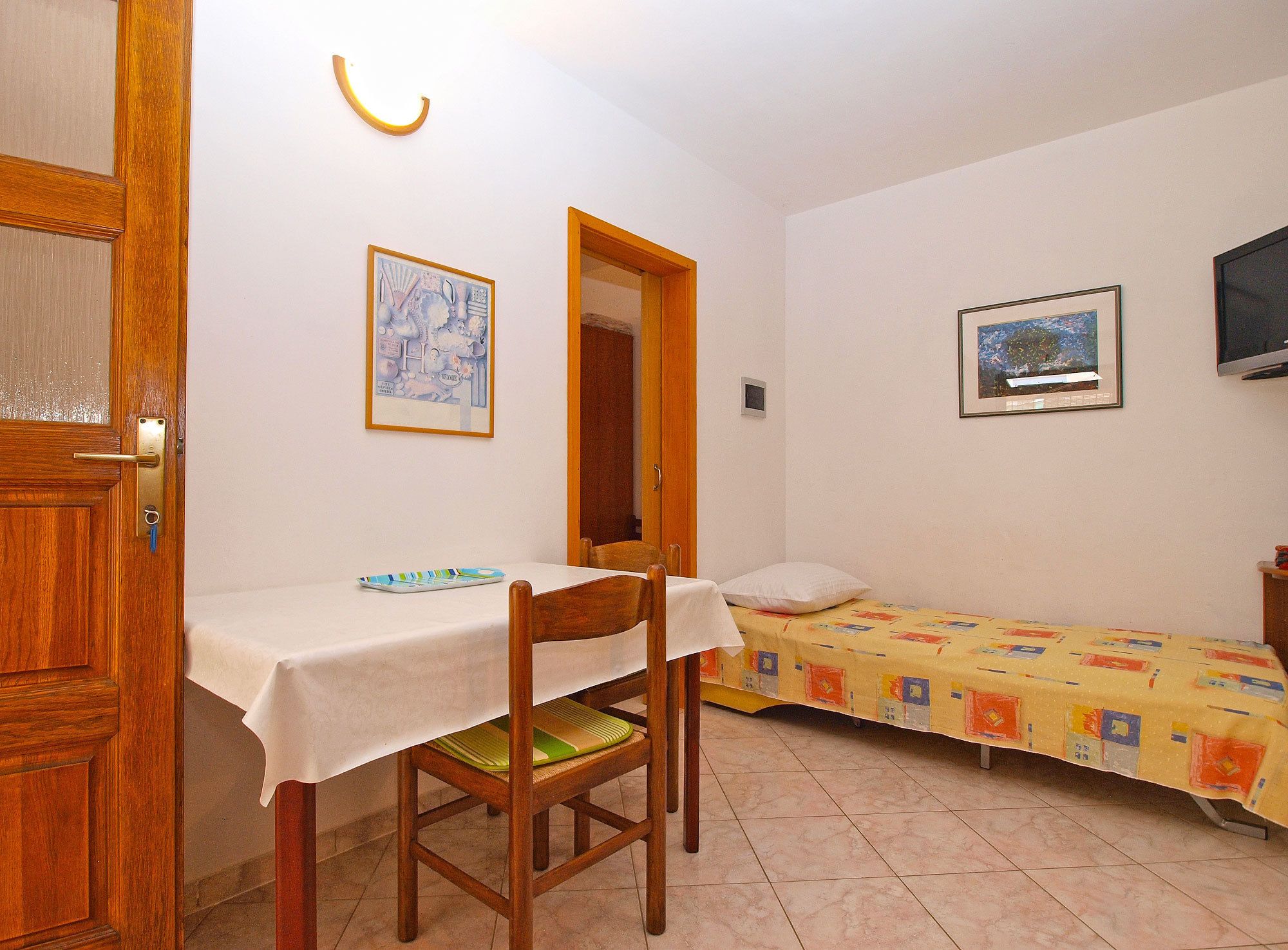 Une petite pièce avec une porte en bois, une table recouverte d’une nappe blanche, une chaise, un lit avec une couvre-lit colorée et deux tableaux encadrés accrochés aux murs.