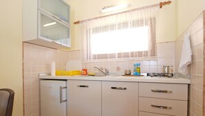 A compact kitchen with a refrigerator, a sink, and a window with lace curtains. - Apartment 1462 (Pula)